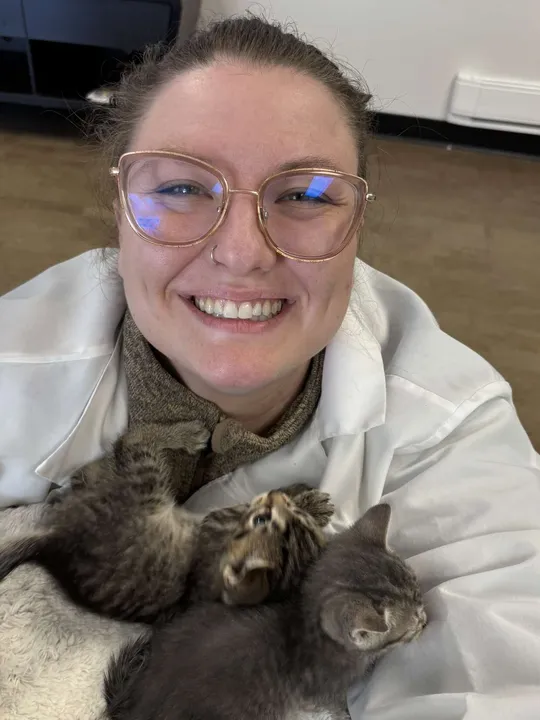 Dr. Edmand smiling excitedly in a selfie-style photo, wearing glasses. Two small kittens are nestled in a blanket on her chest, creating an adorable, heartwarming moment.