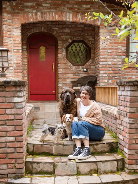 Mel sitting on a stoop in front of a red door with her three dogs, one a large brindle mastiff, the second a blenheim cavalier king charles, and the third is a white and black morkie.