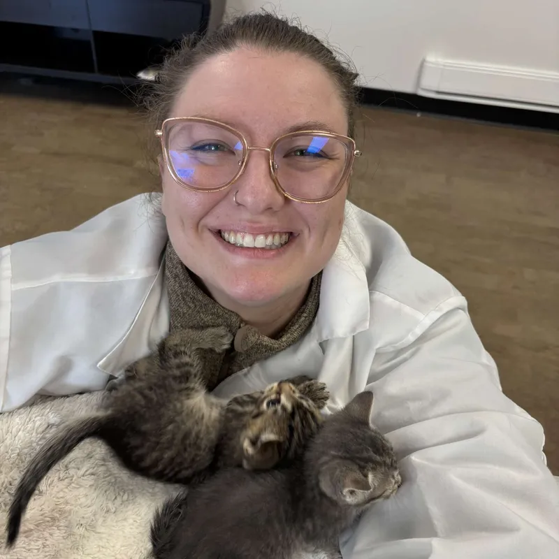 Dr. Edmand smiling excitedly in a selfie-style photo, wearing glasses. Two small kittens are nestled in a blanket on her chest, creating an adorable, heartwarming moment.